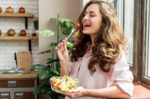 A woman in a pink shirt joyfully eating a salad by a window